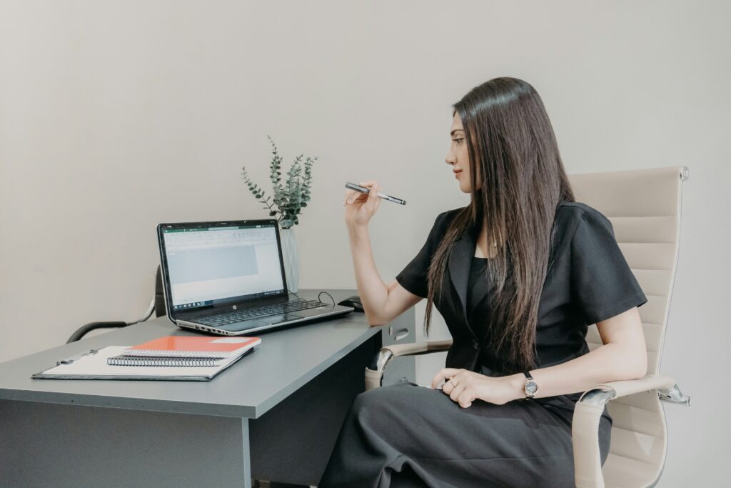 Woman working at desk with laptop and notebooks, holding a pen, focused on screen