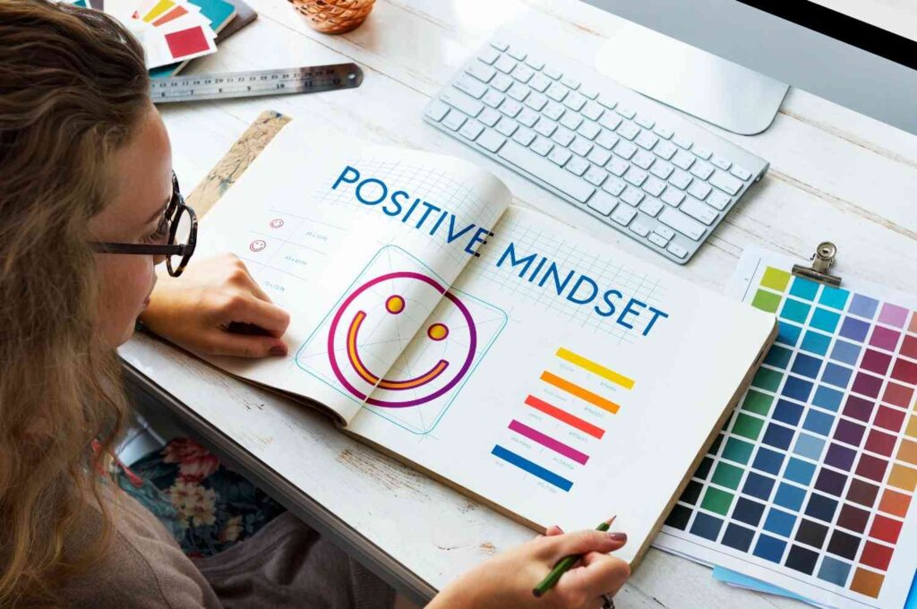 Person reviewing a design notebook with a colorful smiley graphic and the words “Positive Mindset” while working at a desk with a keyboard and color palette.