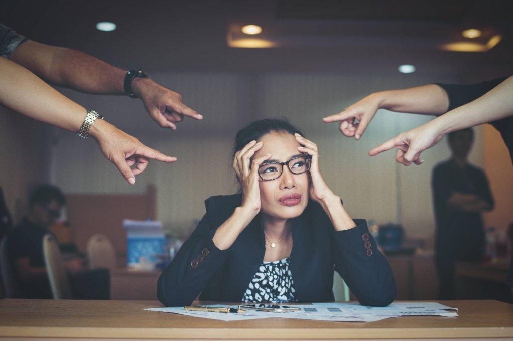 Stressed woman at desk feeling overwhelmed while others point at her representing negative thinking patterns and anxiety